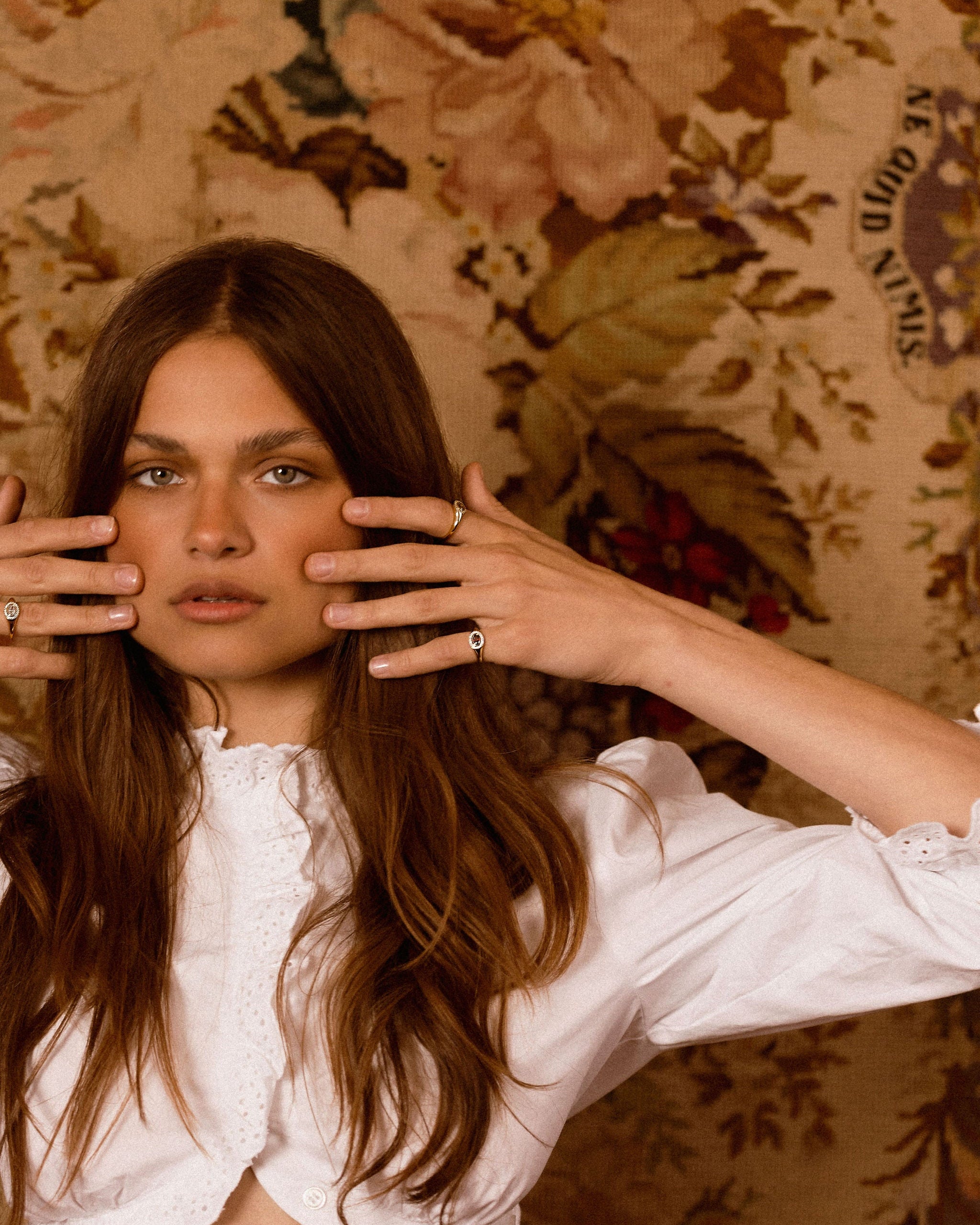 A woman with long brown hair poses in front of a vintage floral wallpaper, wearing a white ruffled blouse and holding her hands near her face.