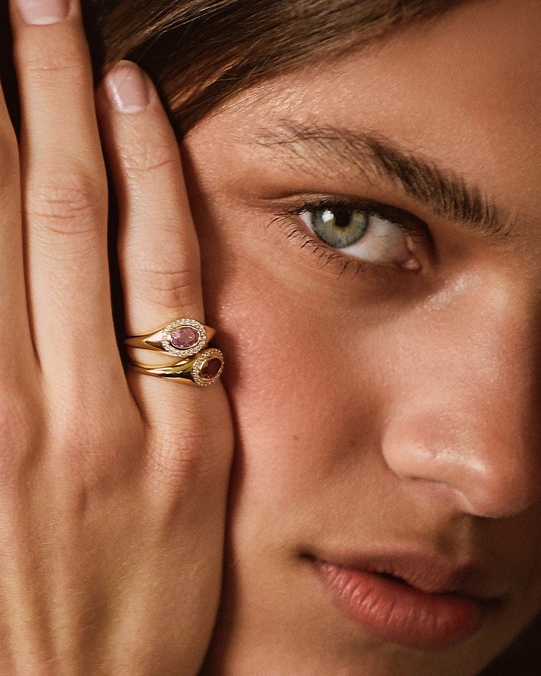 A close-up lifestyle shot of a model wearing a gold ring with a pink gemstone on her finger, with her hand resting against her face.