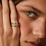 A close-up lifestyle shot of a model wearing a gold ring with a pink gemstone on her finger, with her hand resting against her face.