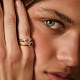 A close-up lifestyle shot of a model wearing a gold ring with a pink gemstone on her finger, with her hand resting against her face.
