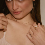 A close-up lifestyle shot of a woman wearing delicate gold jewelry, including a thin gold chain necklace with a small central charm and gold rings on her fingers, paired with a light beige lace camisole.