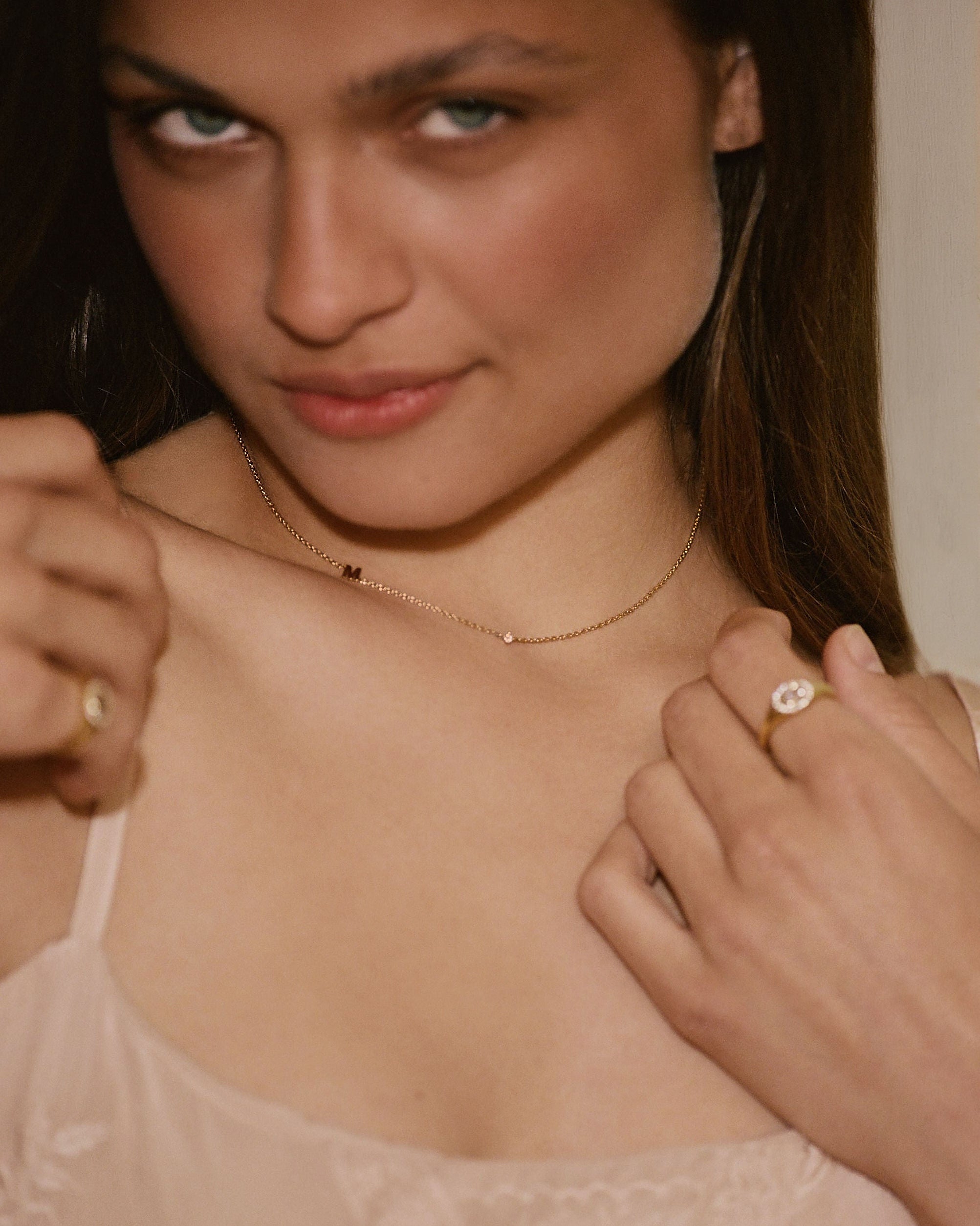 A close-up lifestyle shot of a woman wearing delicate gold jewelry, including a thin gold chain necklace with a small central charm and gold rings on her fingers, paired with a light beige lace camisole.
