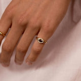 A close-up of a person's hand wearing two small oval rings with gold-toned bands and dark stone centers, resting against light pink fabric.