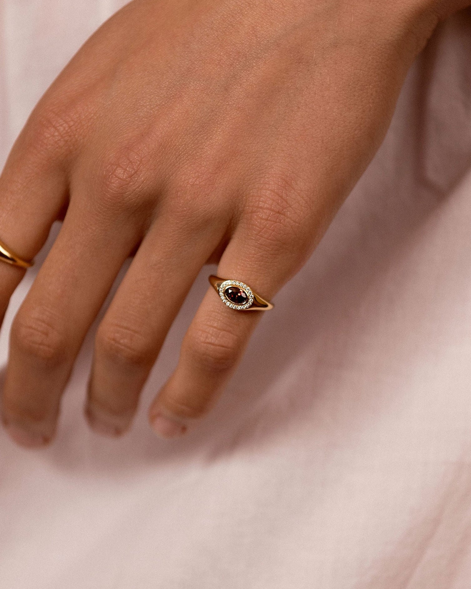 A close-up of a person's hand wearing two small oval rings with gold-toned bands and dark stone centers, resting against light pink fabric.