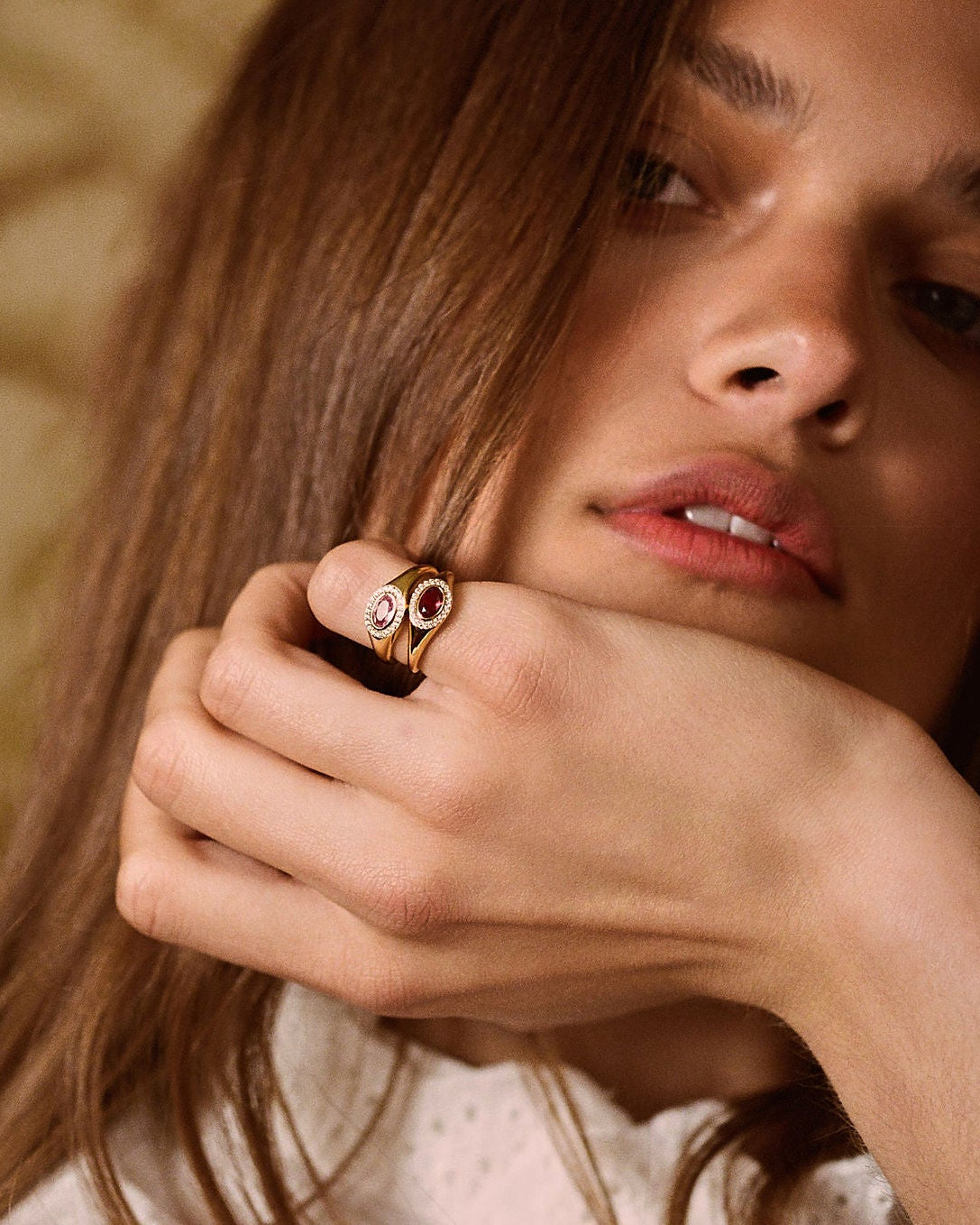 A close-up lifestyle shot of a woman with long brown hair wearing a gold ring with a pinkish-red stone on her finger, set against a warm, patterned background.