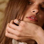 A close-up lifestyle shot of a woman with long brown hair wearing a gold ring with a pinkish-red stone on her finger, set against a warm, patterned background.