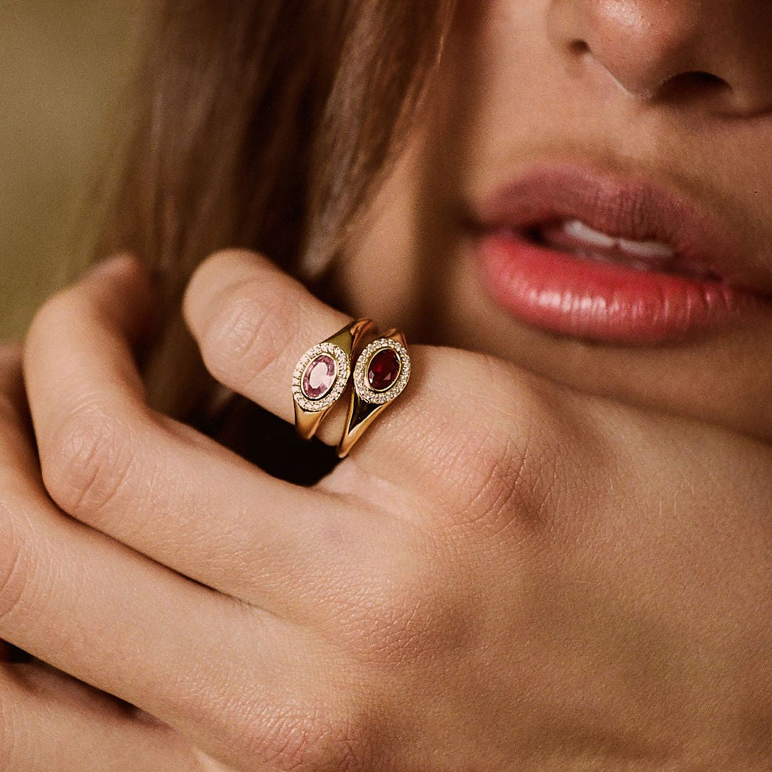 A close-up lifestyle shot of a woman's hand and lower face, showcasing a gold ring with two oval pink gemstones on her ring finger against a warm, blurred brown background.