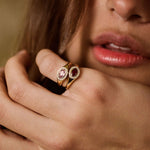 A close-up lifestyle shot of a woman's hand and lower face, showcasing a gold ring with two oval pink gemstones on her ring finger against a warm, blurred brown background.