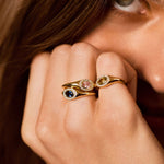 A close-up lifestyle shot of a model's hand wearing two gold rings with colorful gemstones, set against her face and brown hair.