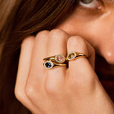 A close-up lifestyle shot of a model's hand wearing two gold rings with colorful gemstones, set against her face and brown hair.