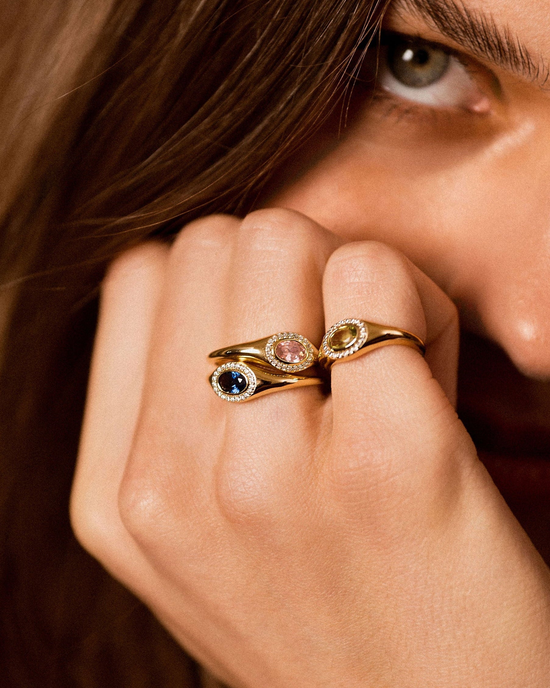 A close-up lifestyle shot of a model's hand wearing two gold rings with colorful gemstones, set against her face and brown hair.