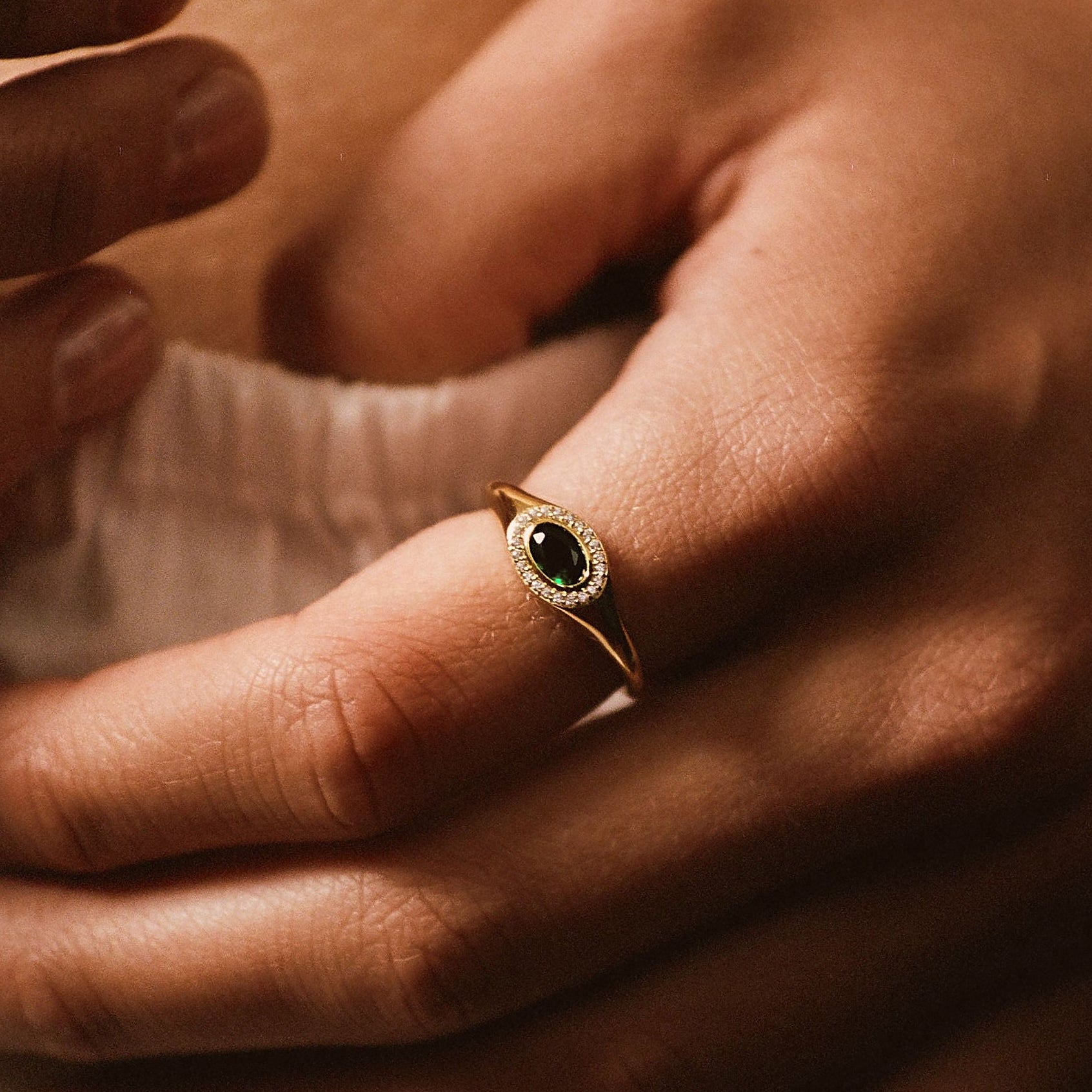 A close-up lifestyle shot of a delicate gold ring with a dark oval stone, worn on a finger of a person's hand resting on their lap.
