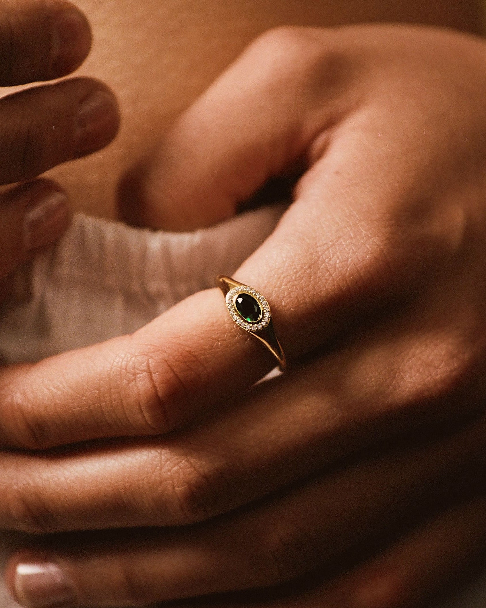 A close-up lifestyle shot of a delicate gold ring with a dark oval stone, worn on a finger of a person's hand resting on their lap.