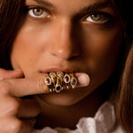 A close-up portrait of a woman holding a cluster of ornate gold rings on her finger, wearing a white lace-trimmed blouse against a dark background.