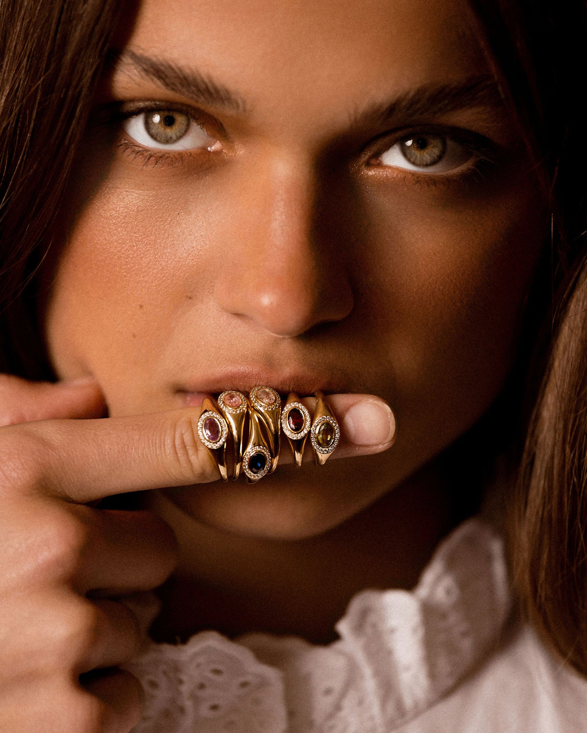 A close-up portrait of a woman holding a cluster of ornate gold rings on her finger, wearing a white lace-trimmed blouse against a dark background.