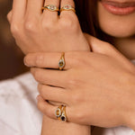 A close-up lifestyle shot of a model's hands wearing multiple gold rings with oval-cut stones, including a green stone and a dark red stone.