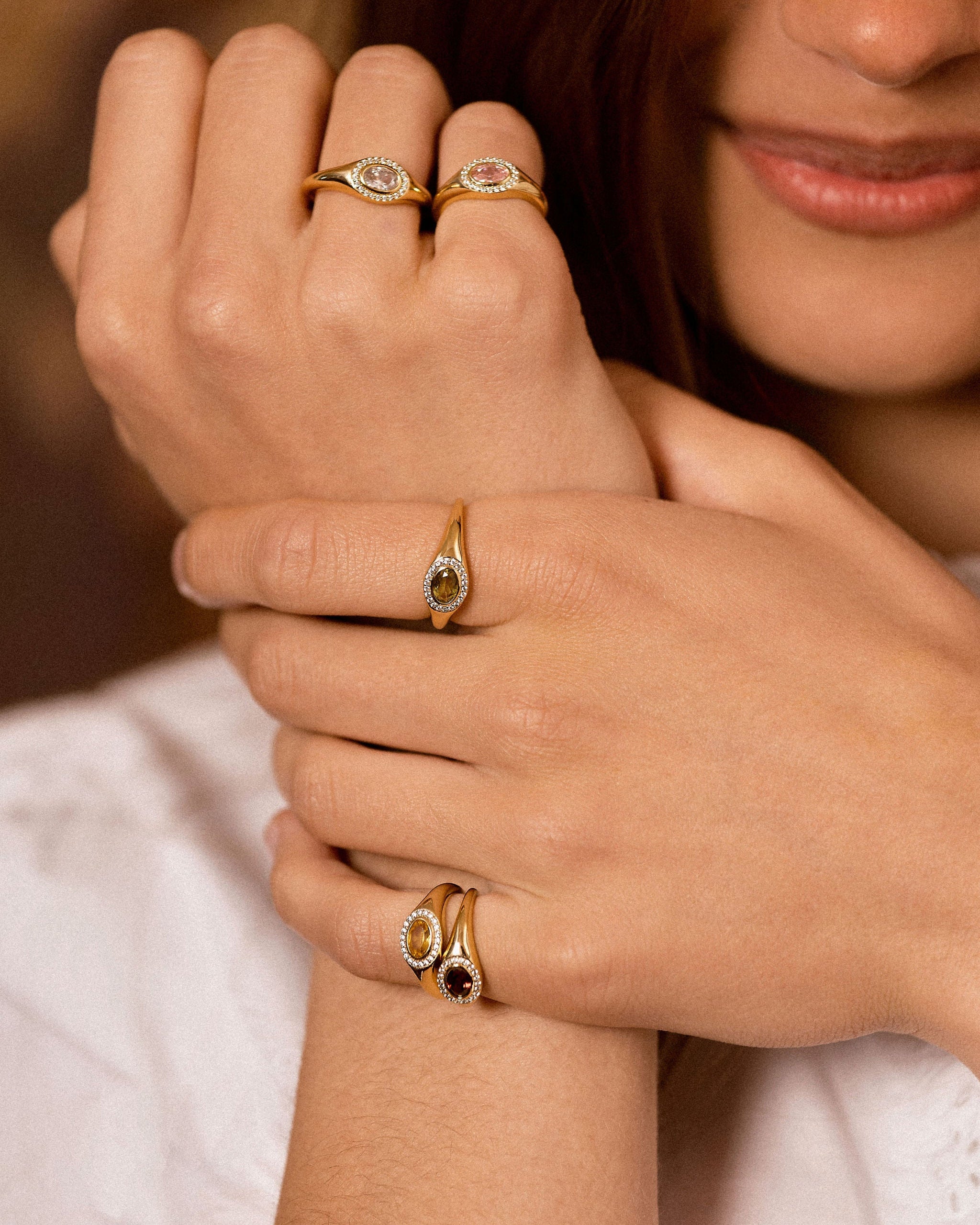 A close-up lifestyle shot of a model's hands wearing multiple gold rings with oval-cut stones, including a green stone and a dark red stone.