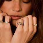 Close-up of a woman resting her chin on her hands, wearing multiple gold rings, including a prominent ring with a red oval gemstone and another with a white stone.