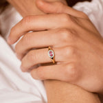 A close-up lifestyle shot of a model wearing a gold ring with a pink oval gemstone on her ring finger, with other gold rings on her other fingers.