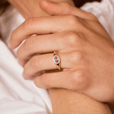 A close-up lifestyle shot of a model wearing a gold ring with a pink oval gemstone on her ring finger, with other gold rings on her other fingers.