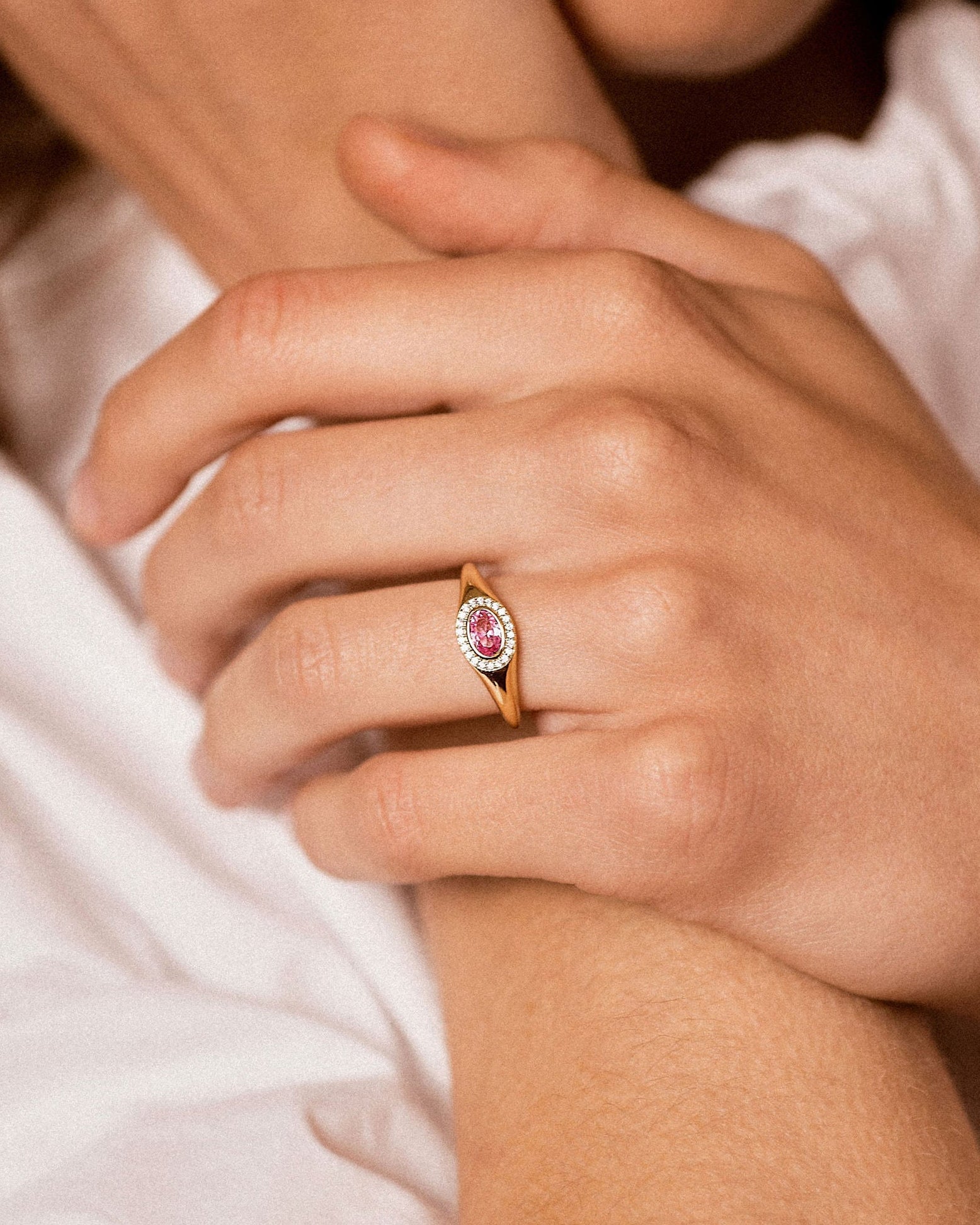 A close-up lifestyle shot of a model wearing a gold ring with a pink oval gemstone on her ring finger, with other gold rings on her other fingers.
