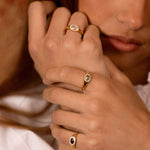 A close-up lifestyle shot of a model wearing three gold rings with oval-cut stones in different colors (clear, light green, and dark red) on her fingers.