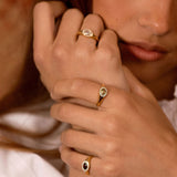 A close-up lifestyle shot of a model wearing three gold rings with oval-cut stones in different colors (clear, light green, and dark red) on her fingers.