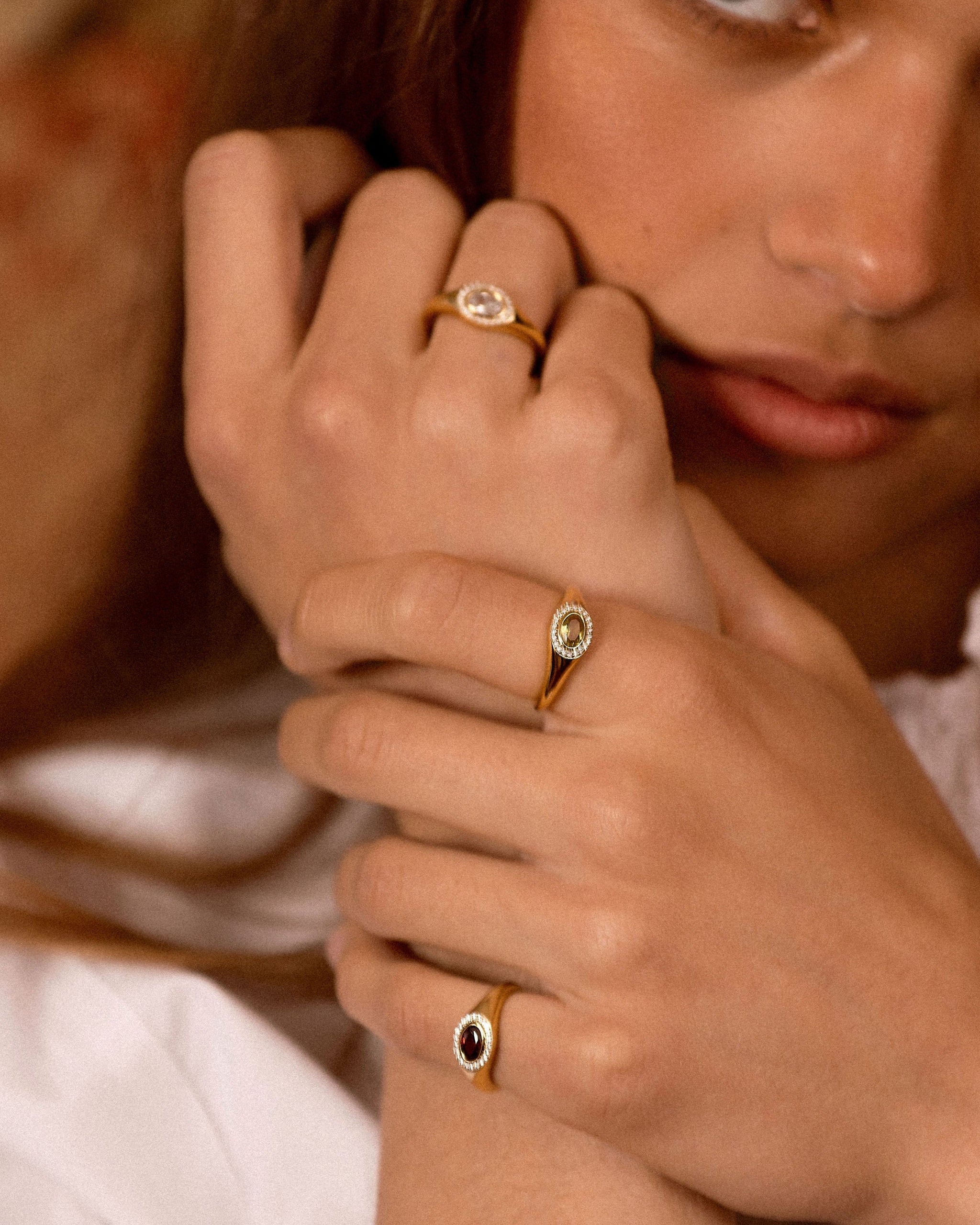 A close-up lifestyle shot of a model wearing three gold rings with oval-cut stones in different colors (clear, light green, and dark red) on her fingers.