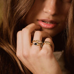 A close-up lifestyle shot of a woman with long brown hair wearing two gold rings on her fingers. One ring features a dark blue oval stone, and the other has a pink oval stone, both set in gold bands.