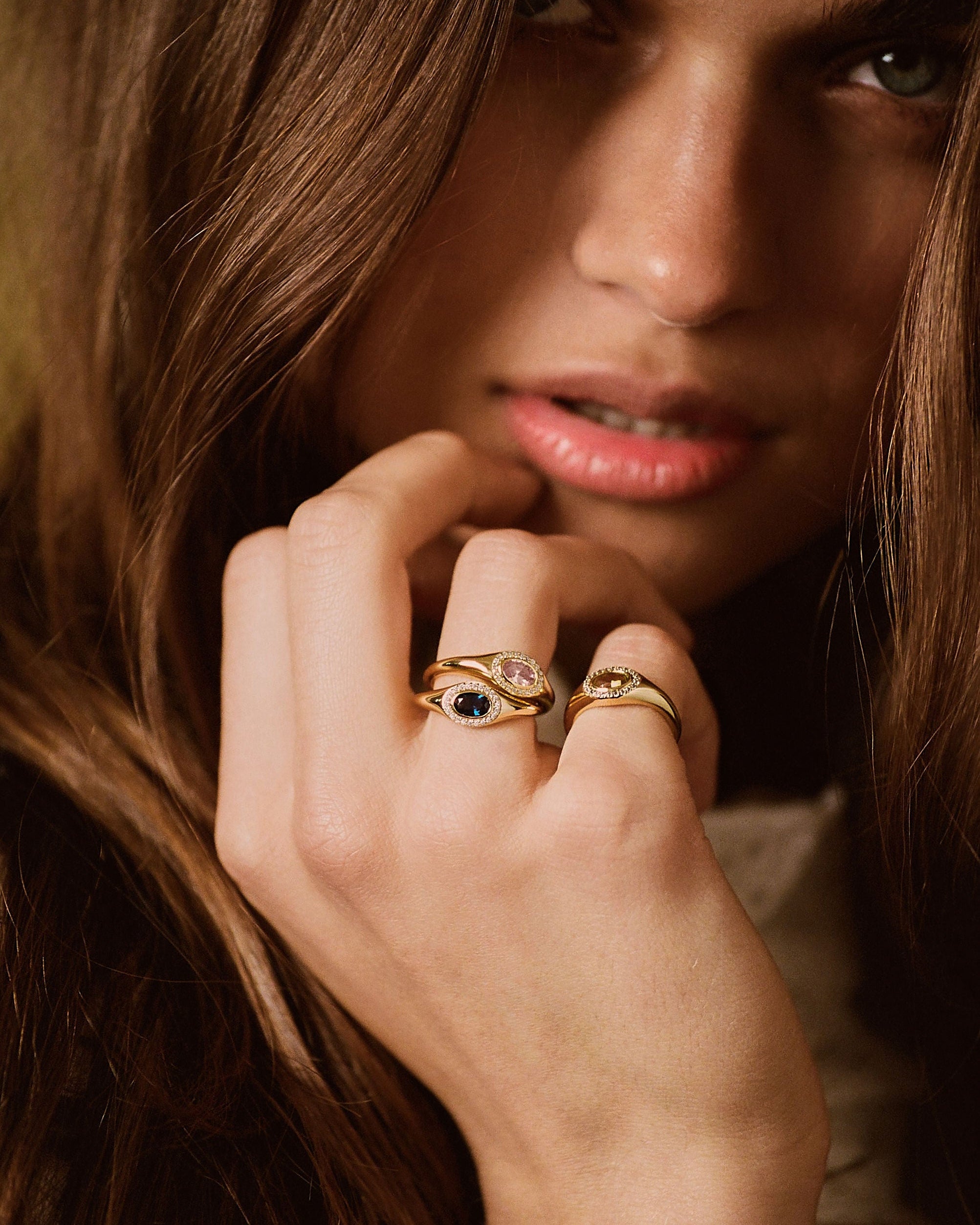A close-up lifestyle shot of a woman with long brown hair wearing two gold rings on her fingers. One ring features a dark blue oval stone, and the other has a pink oval stone, both set in gold bands.