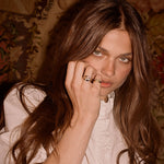 A close-up portrait of a woman with long brown hair wearing multiple gold rings on her fingers, resting her hand near her face against a vintage floral patterned background.