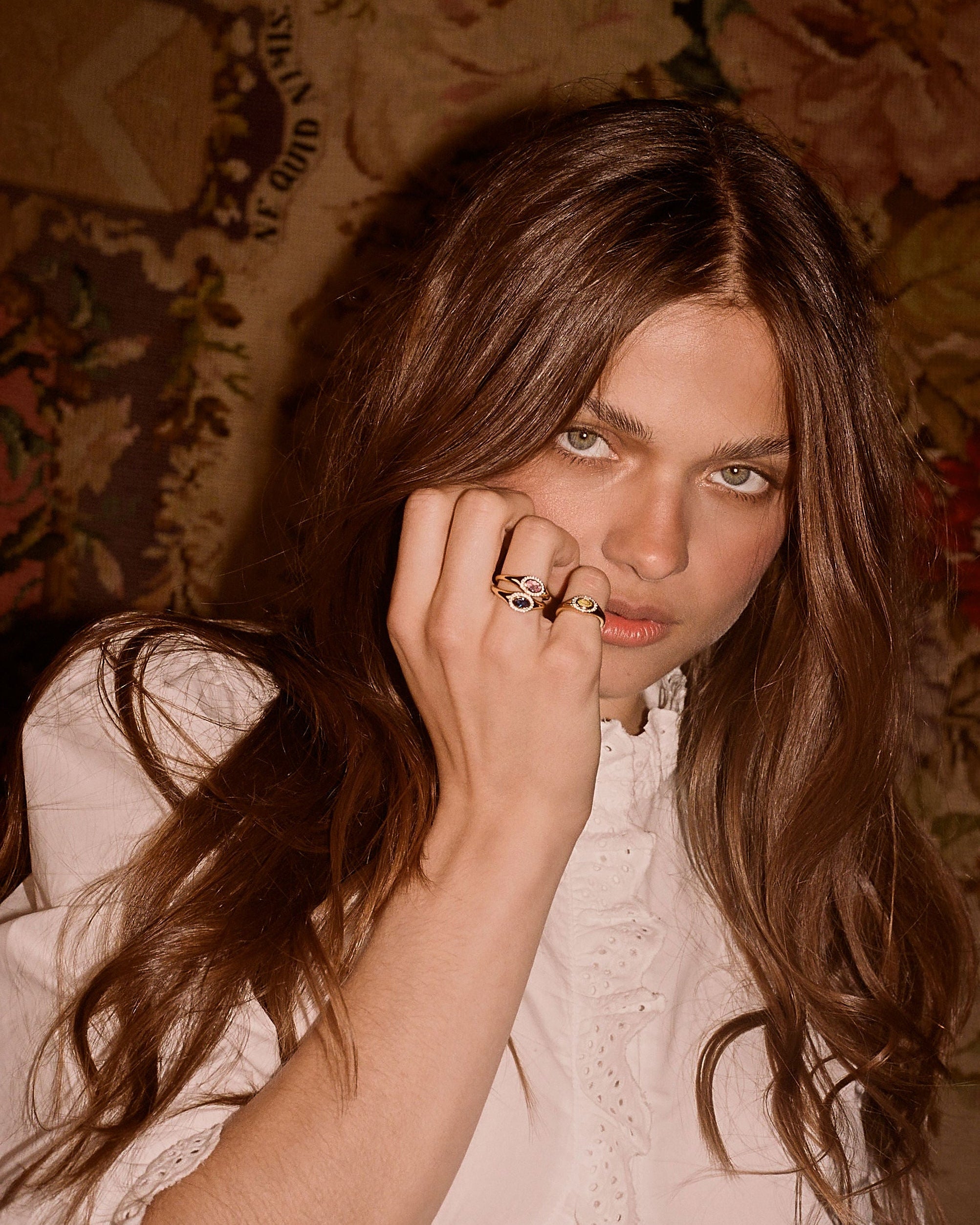 A close-up portrait of a woman with long brown hair wearing multiple gold rings on her fingers, resting her hand near her face against a vintage floral patterned background.