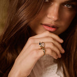 A close-up lifestyle shot of a woman with long brown hair wearing a gold ring with a double oval-cut stone setting on her finger.
