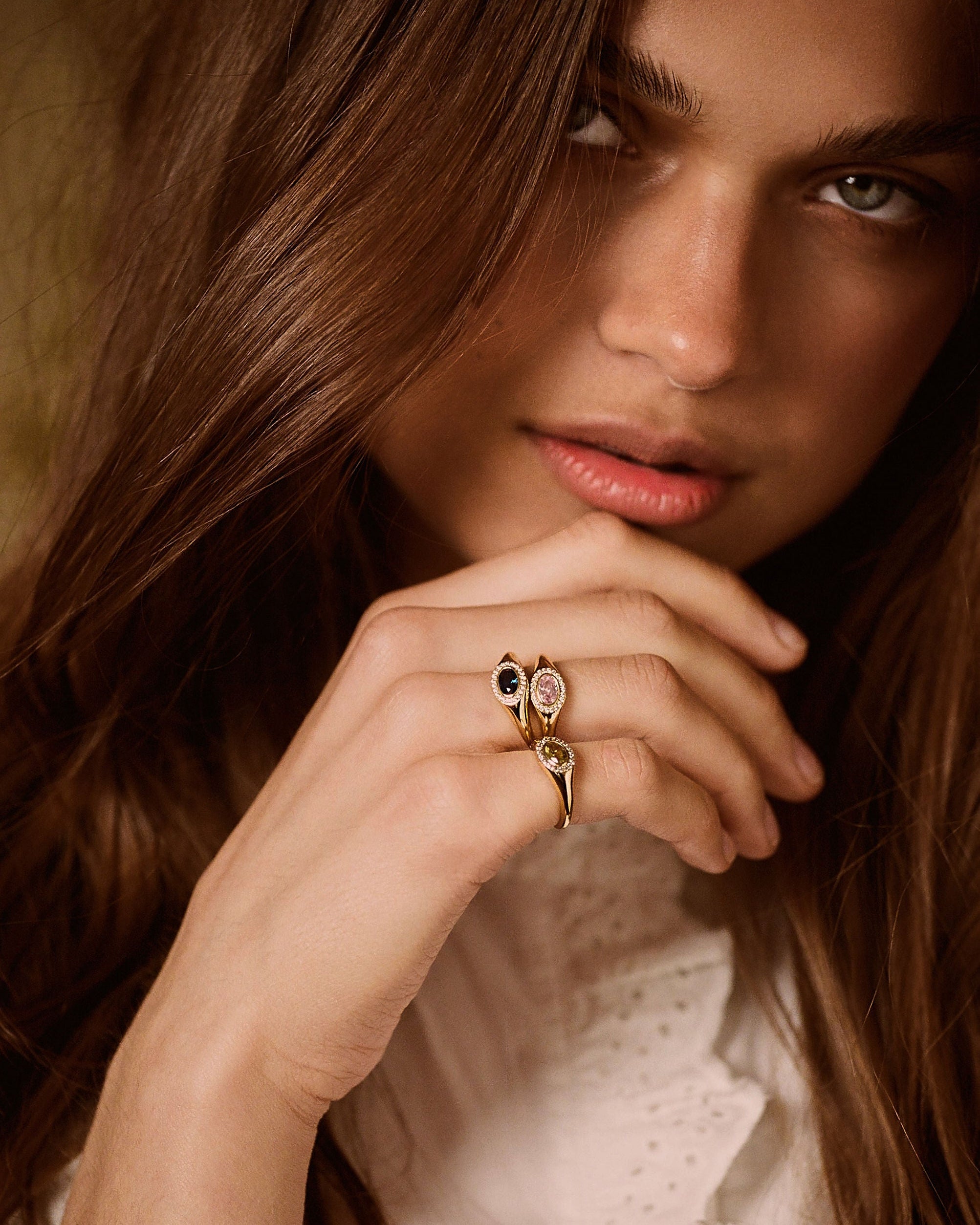 A close-up lifestyle shot of a woman with long brown hair wearing a gold ring with a double oval-cut stone setting on her finger.