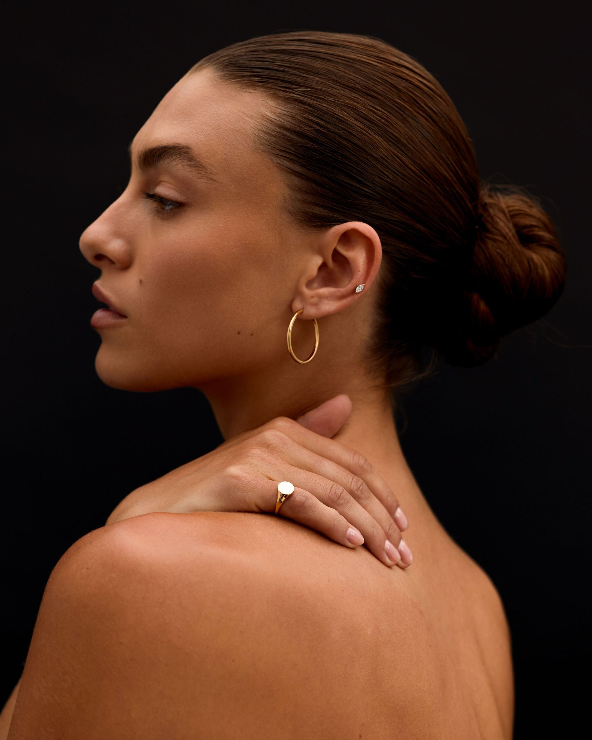 Woman with a bun, wearing gold earrings and a ring, against a black background
