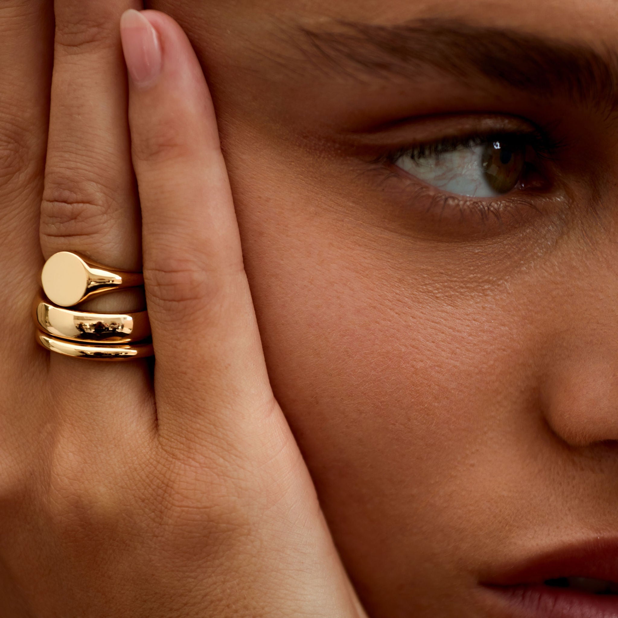 Close-up of a person wearing a gold ring on their finger, with a hand covering part of the face.