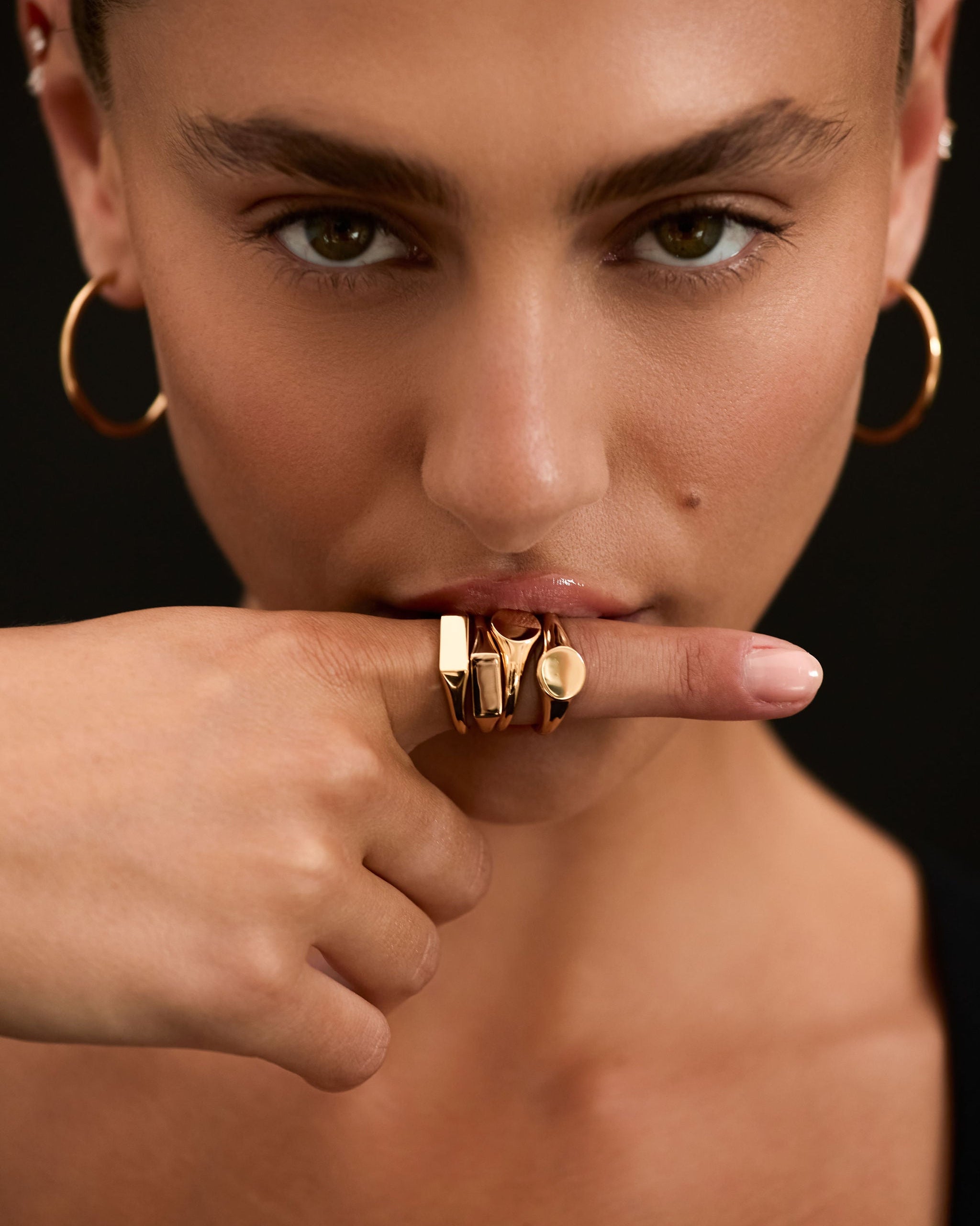 Woman wearing gold hoop earrings and rings on a black background