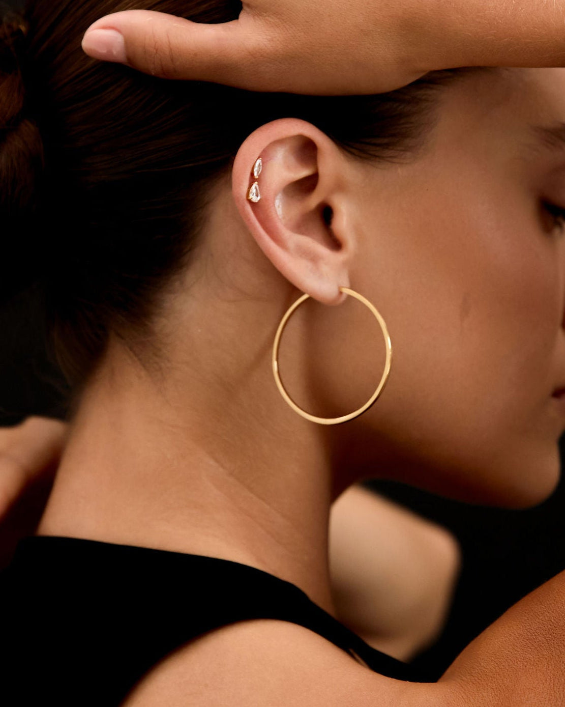 Woman with gold hoop earrings and watch adjusting her hair against a black background