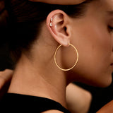 Woman with gold hoop earrings and watch adjusting her hair against a black background