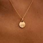 Gold necklace with a round pendant on a person's neck against a brown background