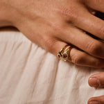 A close-up of a hand wearing a gold ring with a small pink gemstone, resting on a light beige fabric surface.