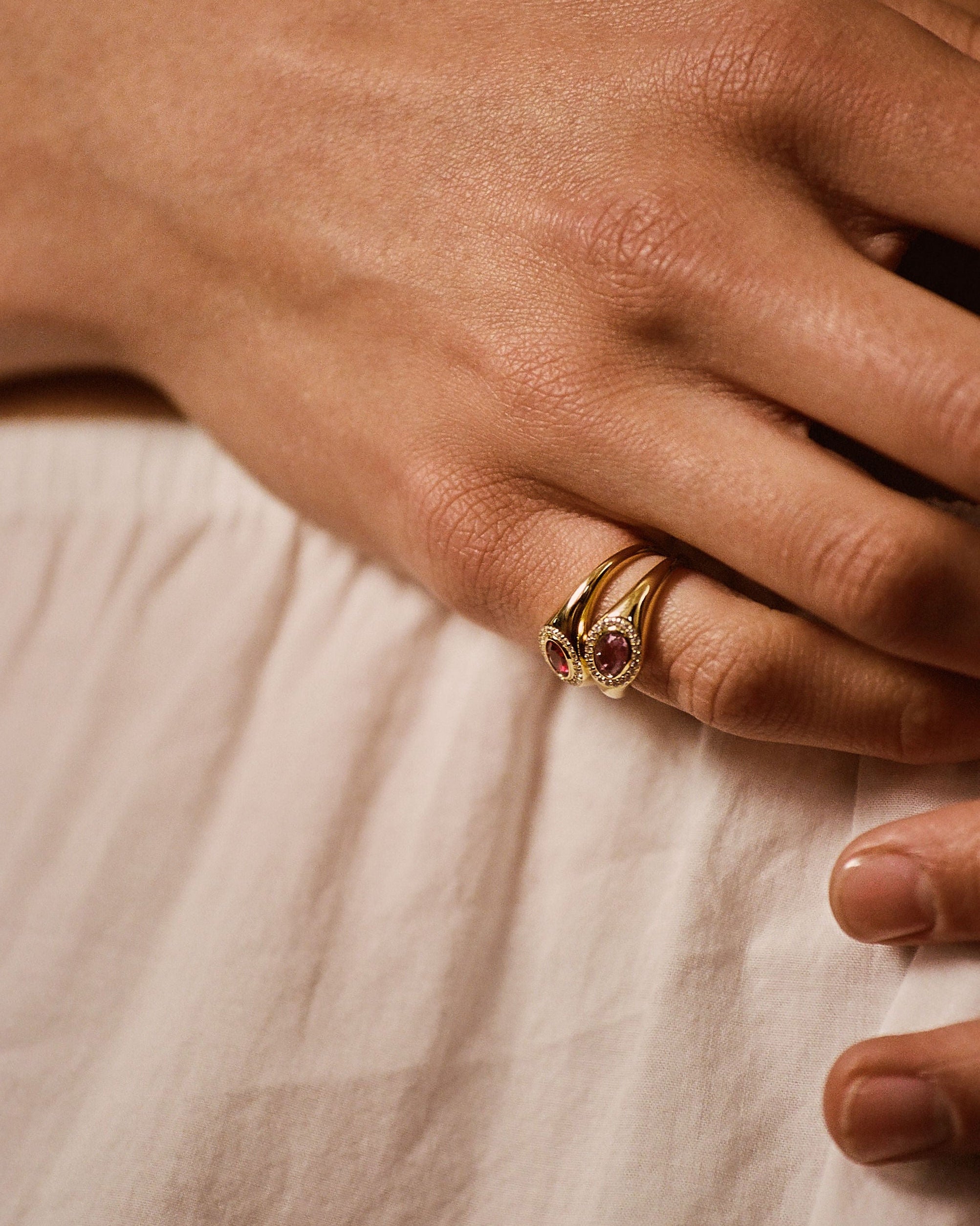 A close-up of a hand wearing a gold ring with a small pink gemstone, resting on a light beige fabric surface.